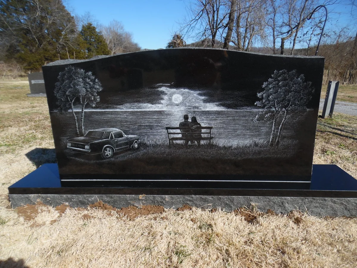 An engraving on black granite shows two people on a bench beside water, with a moon, trees and a car