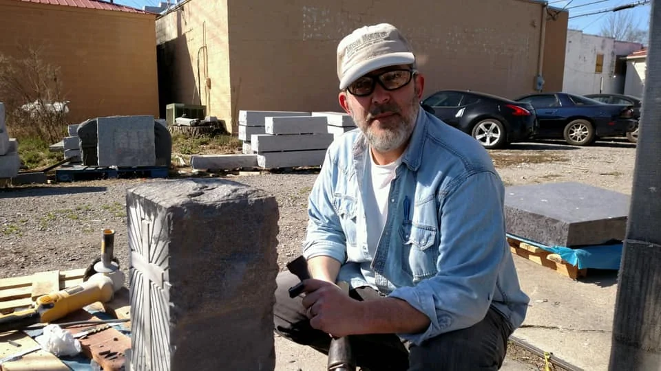 A man wearing a cap and glasses beside a piece of stone and hand tools
