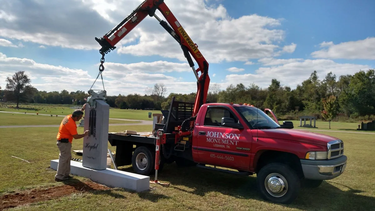 A person in an orange shirt working beside a tall memorial in the cemetery, with the red truck’s boom still attached to the stone