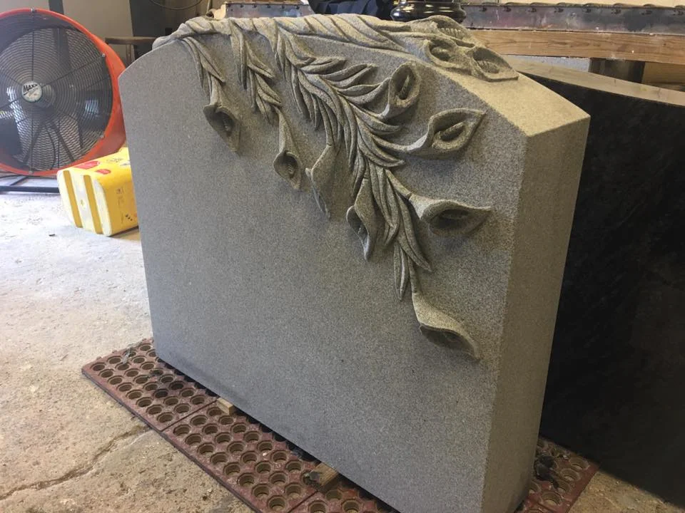 Deeply sculpted flowers and leaves curve over the top and side of a grey granite memorial in the workshop