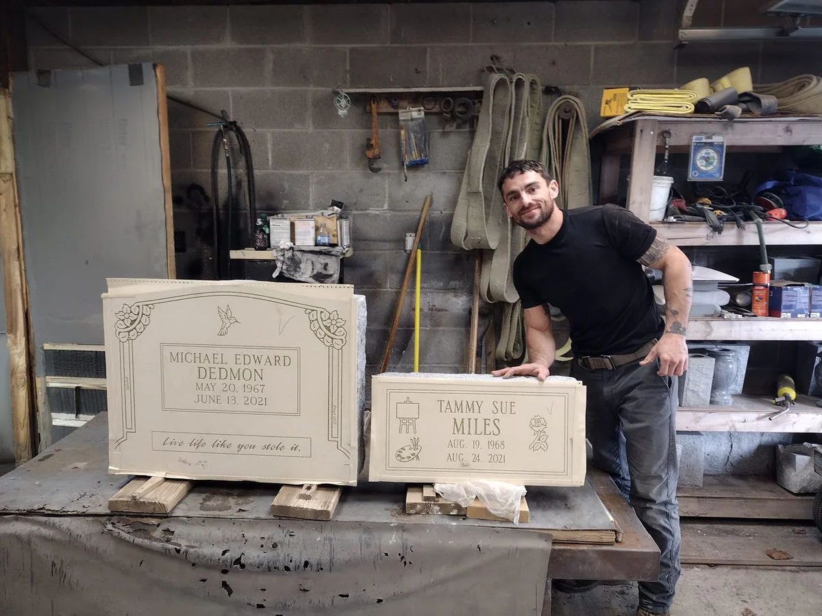 A Johnson Monument Company craftsperson stands beside two prepared memorial faces on a workshop bench